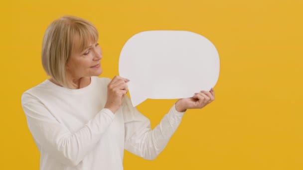 Good looking senior lady holding blank speech bubble with free space and smiling to camera, orange studio background - Záběry, video