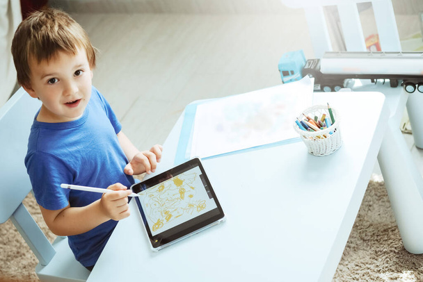 Lindo chico en camiseta azul usando tableta digital o computadora para dibujar su imagen, usando lápiz. Arte de las nuevas tecnologías, educación en línea - Foto, Imagen
