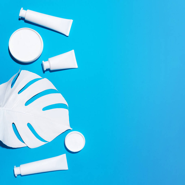Cosmetics, skin care, beauty, body treatment concept. White cosmetic jar, tube, bottle and tropical monstera leaf over blue background. Top view. Flat lay. Mock-up. - Fotó, kép