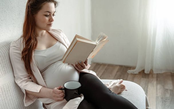 Stilvolle schwangere Frau in den letzten Monaten der Schwangerschaft ruht zu Hause mit einem Buch und einem Drink in einer Tasse. - Foto, Bild