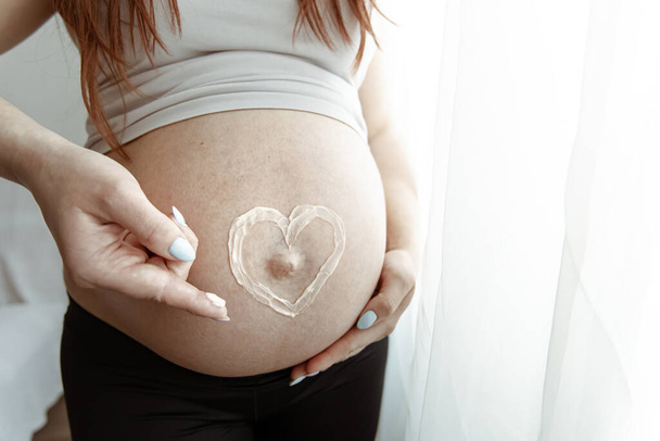Nahaufnahme des nackten Bauches einer zukünftigen Mutter in den letzten Monaten der Schwangerschaft mit einer bemalten Herzcreme. - Foto, Bild