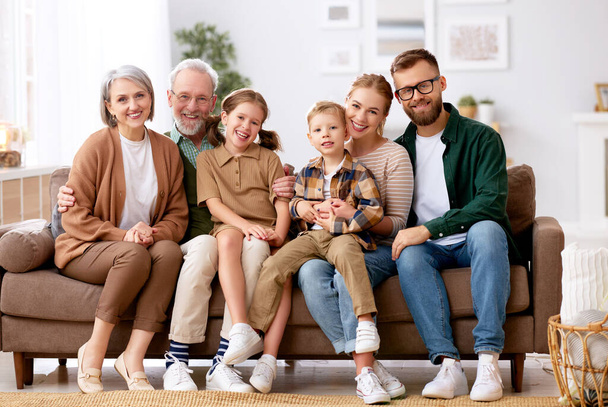 Big happy family. Portrait of grandparents, mother, father and two their cute kids, sister and brother, sitting together on coach at home and smiling at camera. Mortgage loan and real estate concept - Photo, Image