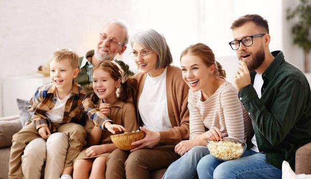 Happy big family enjoying weekend together at home. Grandparents, mother, father and two little kids, sister and brother, sitting on coach and eating popcorn while watching funny movie comedy on TV - Photo, Image
