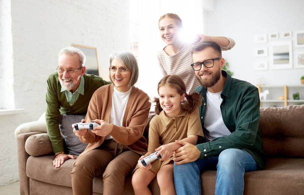 Schöne große Mehrgenerationenfamilie, die das Wochenende zusammen zu Hause verbringt. Aufgeregte Großeltern, Mutter und Vater mit kleinen Kindern spielen Videospiele und amüsieren sich auf dem Sofa im Wohnzimmer - Foto, Bild