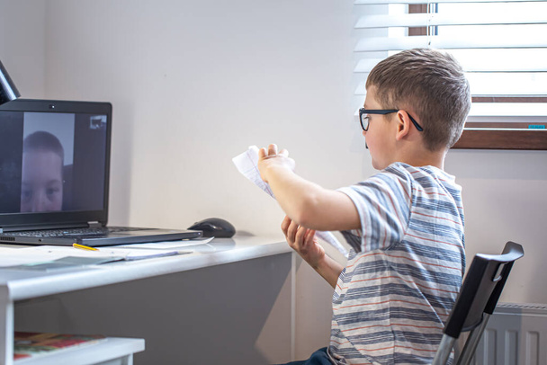 Een basisschoolstudent zit achter een bureau voor een laptop en communiceert thuis via videolink online. - Foto, afbeelding