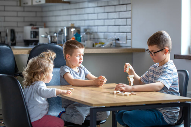 Small children play a board game with wooden cubes at home in the kitchen. - Фото, изображение