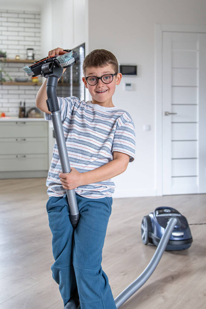 Egy szemüveges kisfiú szórakozik és porszívóval takarítja a házat.. - Fotó, kép