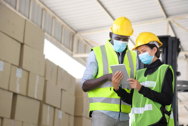 Les travailleurs afro-américains et asiatiques portant un masque facial et un gilet de sécurité travaillant dans un entrepôt vérifiant l'inventaire à l'aide d'une tablette numérique pendant la nouvelle normale après covid-19 - Photo, image