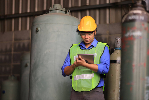 Digitális táblagépet használó ázsiai ipari dolgozó a hűtőfolyadék-rendszer ellenőrzéséhez a gyárban, miközben folyékony nitrogén és argon előtt áll a biokémiai ipar sűrített tartályában - Fotó, kép