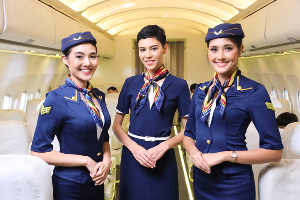 Joukkue erilaisia lentoemäntä poseeraa hymyillen keskellä käytävää sisällä ilma-aluksen matkustajat istuvat taustalla - Valokuva, kuva