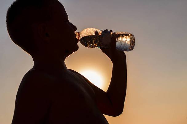 Ένα αγόρι με κοντά μαλλιά πίνει νερό από ένα μπουκάλι σε ένα αντικριστό φως. σιλουέτα - Φωτογραφία, εικόνα