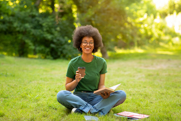 Cheerful black girl having rest, drinking coffee and reading book at campus - Photo, Image