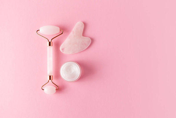 Rose quartz facial roller, gua sha and moisture cream for skincare, top view. Trendy skincare facial massager tools on pink background - 写真・画像