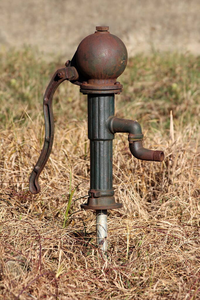 Strange and unusually shaped vintage retro partially rusted wrought iron dark green hand water pump mounted on grey water pipe surrounded with dry grass in suburban family house backyard on warm sunny winter day - Photo, Image
