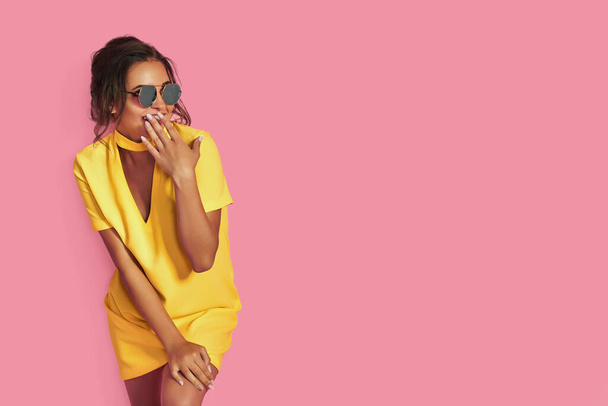 Beautiful girl in yellow jeans and pink shirt, with hands up wearing sunglasses posing, dancing, smiling on pink background in studio. - Foto, Imagem