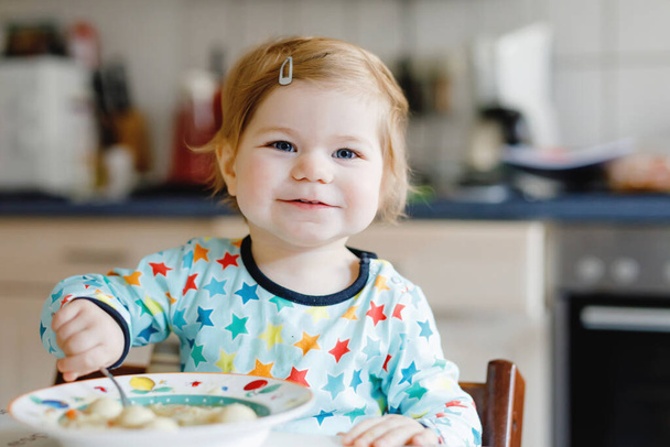 Entzückendes kleines Mädchen, das von einer löffelweise Gemüsenudelsuppe isst. Ernährung, Kind, Ernährung und Entwicklungskonzept. Niedliches Kleinkind, Tochter mit Löffel sitzt im Hochstuhl und lernt selbst zu essen. - Foto, Bild