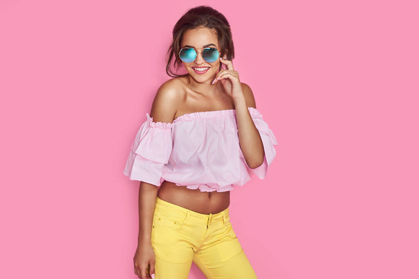 Beautiful girl in yellow jeans and pink shirt, with hands up wearing sunglasses posing, dancing, smiling on pink background in studio. - Photo, image