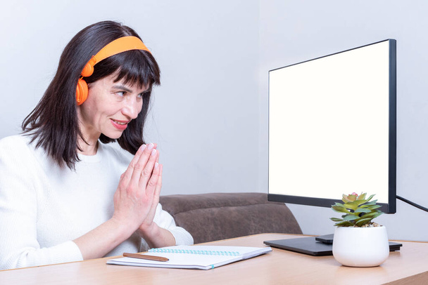 Smiling business woman in headphones looks at a monitor with a blank white screen and shows a sign of thanks, gratitude, clasped palms together, side view, copy space. Business online concept - Zdjęcie, obraz