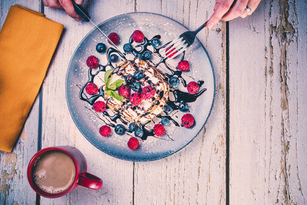 top view of a person's hands with knife and fork to eat a delicious breakfast of pancakes with fresh fruits and chocolate syrup and a hot coffee. - Fotó, kép