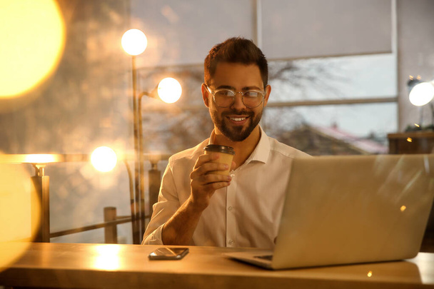 Человек с чашкой кофе работает на ноутбуке за столом в офисе - Фото, изображение
