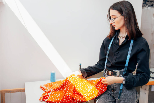 Side view of a fashion designer looking at new fabric for an online store. Young woman business owner sitting on the desk and working with a garment for a new collection in her creative office.  - Foto, afbeelding