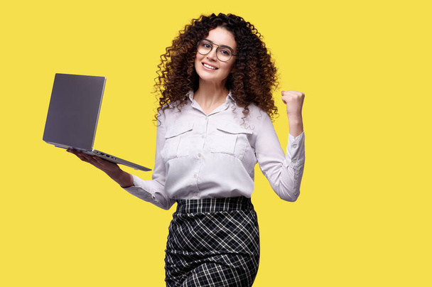Mulher feliz em camisa branca e óculos com laptop se alegra e segura punho no fundo amarelo isolado - Foto, Imagem