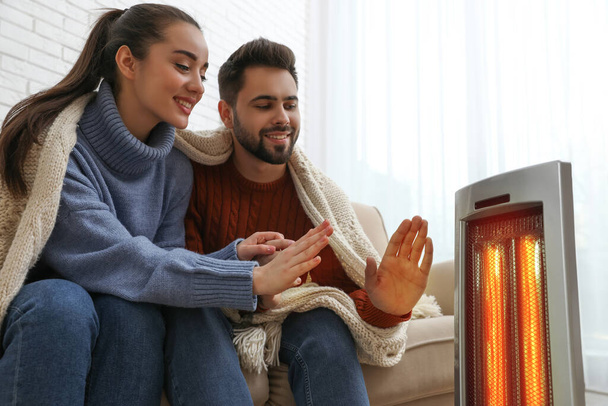 Young couple warming hands near electric heater at home - Fotografie, Obrázek