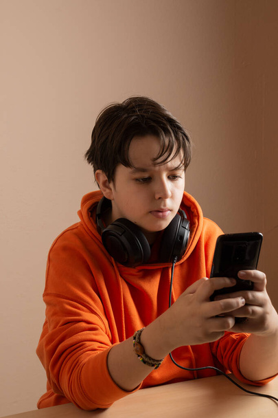 Хлопчик-підліток з помаранчевою сорочкою та навушниками, дивиться на мобільний, з концентрованим обличчям, портрет
 - Фото, зображення