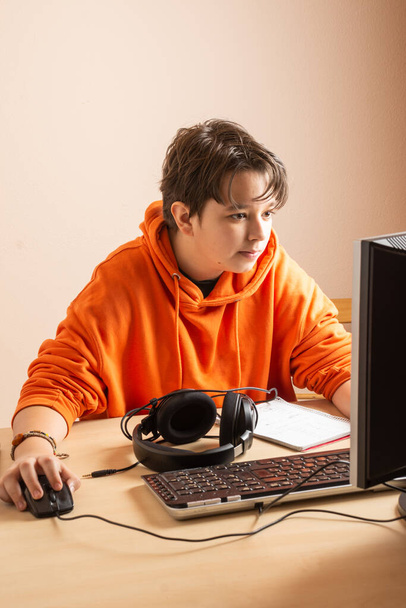 Niño adolescente en sudadera naranja con ordenador en la mesa, mirando a la pantalla con el ratón en la mano, retrato - Foto, Imagen