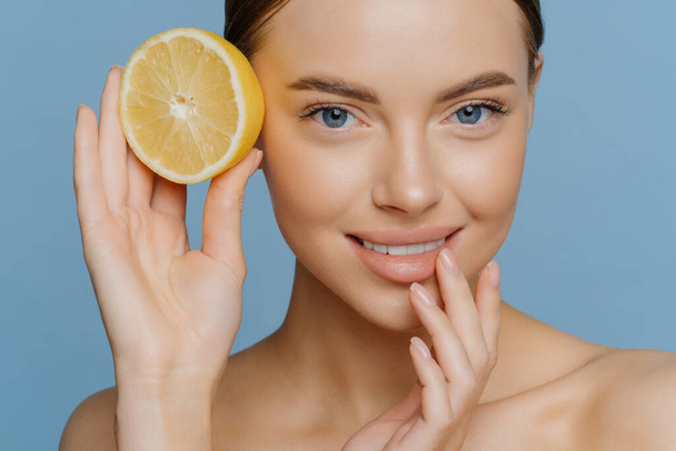 Natural component for face and body care. Close up shot of lovely young European woman smiles gently keeps hand near mouth holds half of lemon stands bare shoulders isolated over blue background - Photo, Image