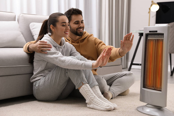 Young couple warming hands near electric heater at home - Fotoğraf, Görsel