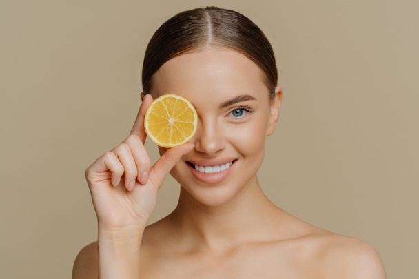 Beauty and vitamin C concept. Headshot of lovely dark haired European woman smiles gently covers eye with slice of juicy lemon poses half naked against brown wall has healthy skin eats healthy food. - Photo, Image