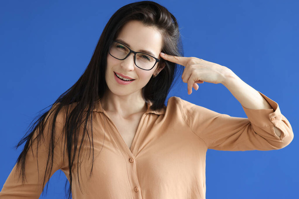 Νεαρή γυναίκα με γυαλιά κρατώντας δείκτη κοντά στο μέτωπο σε μπλε φόντο - Φωτογραφία, εικόνα