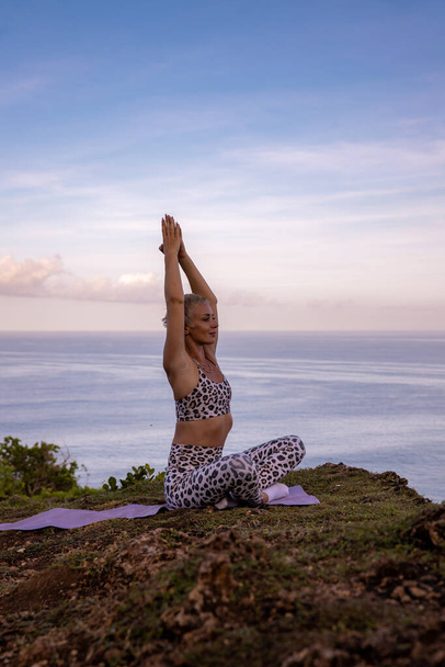 Práctica de yoga al amanecer. Mujer joven practicando Padmasana, Lotus Pose. Asana sentada. Manos levantadas en namaste mudra. Estilo de vida saludable. Retiro de yoga. Acantilado de Uluwatu, Bali, Indonesia - Foto, imagen