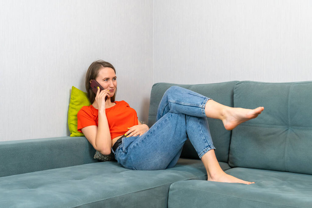 Νεαρή όμορφη μελαχρινή γυναίκα με μπλε τζιν και πορτοκαλί πουκάμισο είναι ξαπλωμένη στον καναπέ και μιλάει στο τηλέφωνο - Φωτογραφία, εικόνα