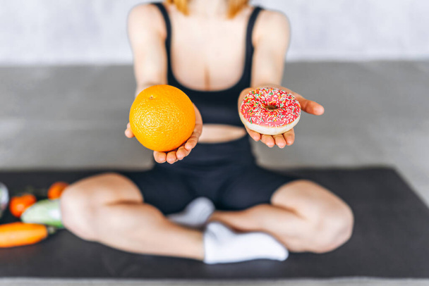 Urheilullinen nainen, jolla on donitsi ja appelsiini käsissä. Terveellisen ja epäterveellisen ruoan käsite. - Valokuva, kuva