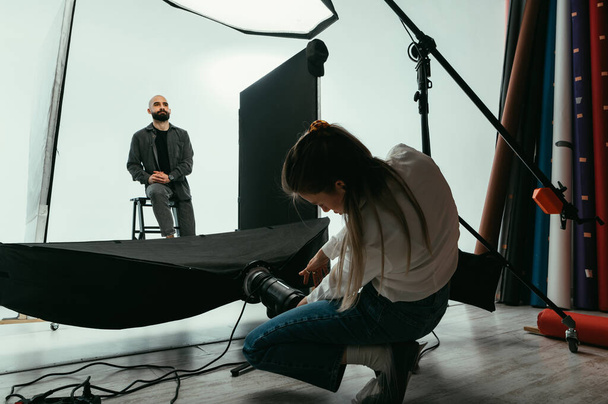 Kobieta fotograf podczas fotografowania modelu dostosowuje dolną lampę błyskową. Sesja zdjęciowa za kulisami dla mężczyzny. - Zdjęcie, obraz
