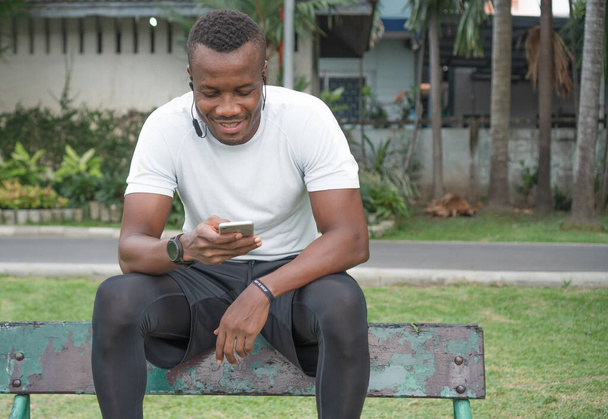 feliz e sorridente homem afro-americano muscular em sportswear com um smartphone ouvindo música em cadeira de madeira no parque. Atleta masculino positivo e saudável relaxar após o treinamento. - Foto, Imagem