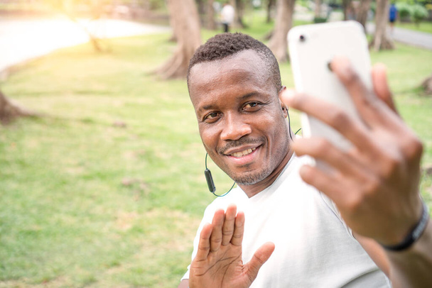mladý Afroameričan mluví v parku na svém chytrém telefonu. Mladý černý sportovec odpočívá po tréninku v zeleném lese. pohledný sportovec selfie a vlog streaming při cvičení. - Fotografie, Obrázek