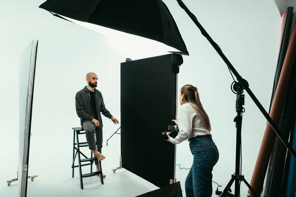 Vrouw fotograaf maakt portretten voor een man met een baard op een witte achtergrond in een fotostudio, communiceert met een model tijdens het fotograferen. - Foto, afbeelding