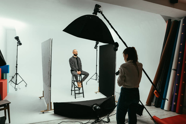 Vrouwenfotograaf maakt portretten voor een knappe jongeman, zit in een stoel op een witte achtergrond en poseert met een glimlach op zijn gezicht. - Foto, afbeelding
