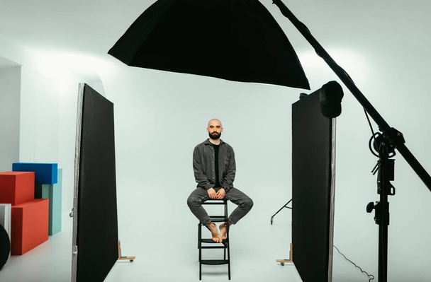 Bärtiges männliches Model sitzt auf einem Stuhl in einem Fotostudio auf weißem Hintergrund und blickt mit ernstem Gesicht in die Kamera. - Foto, Bild
