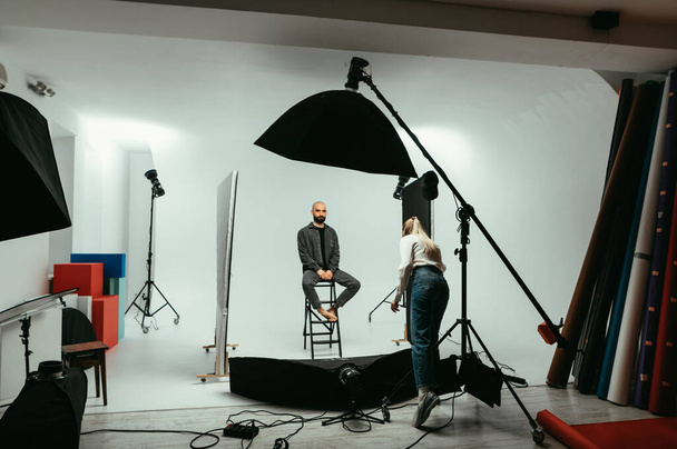 Mulher fotógrafa e modelo homem barbudo trabalhando em um estúdio de fotografia em um fundo branco com flashes. Backstage foto processo sessão de fotos. - Foto, Imagem