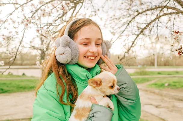 Adolescente em um casaco verde com um filhote de cachorro chihuahua em seus braços na primavera. árvores estão florescendo - Foto, Imagem