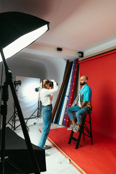 Una fotógrafa con una cámara en las manos en un estudio fotográfico hace una sesión de fotos para un guapo barbudo sobre un fondo rojo. Vertical - Foto, Imagen