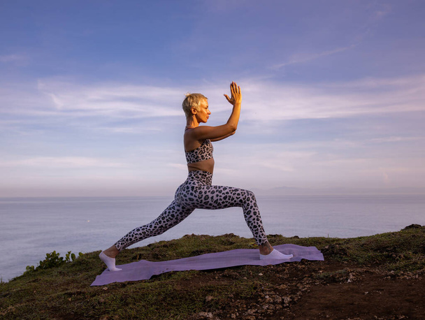 Young woman practicing Virabhadrasana I, Warrior I Pose. Hands in namaste mudra. Yoga retreat. Healthcare concept. Balance and concentration. Healthy lifestyle. Uluwatu cliff, Bali, Indonesia - Photo, Image