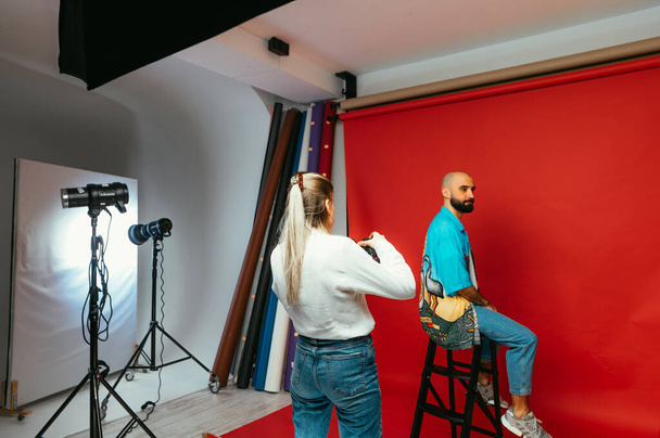Γυναίκα επαγγελματίας φωτογράφος με μια φωτογραφική μηχανή στα χέρια του κάνει μια φωτογράφηση για ένα γενειοφόρο όμορφο άνδρα σε ένα κόκκινο φόντο σε ένα στούντιο φωτογραφιών. - Φωτογραφία, εικόνα