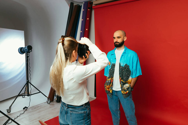 Kobieta fotograf robi portrety dla przystojnego brodatego mężczyzny w studio fotograficznym na czerwonym tle, za kulisami zdjęcie. - Zdjęcie, obraz