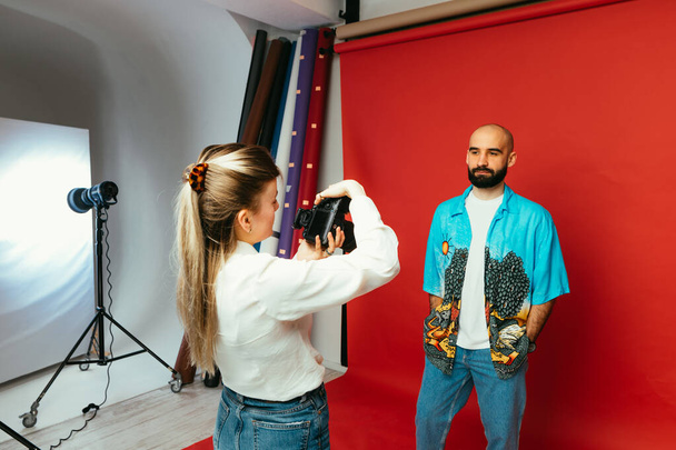 Foto entre bastidores, fotógrafa profesional femenina con una cámara en las manos hace una sesión de fotos para un hombre con ropa de color sobre un fondo rojo en un estudio de fotografía. - Foto, Imagen