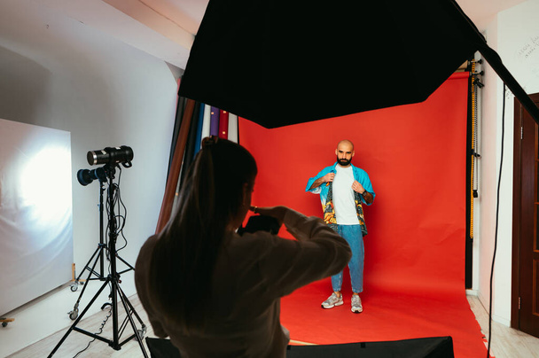 Mulher fotógrafa atira em um homem barbudo em roupas brilhantes em um fundo vermelho em um estúdio de fotos. Conceito de trabalho de fotógrafo - Foto, Imagem